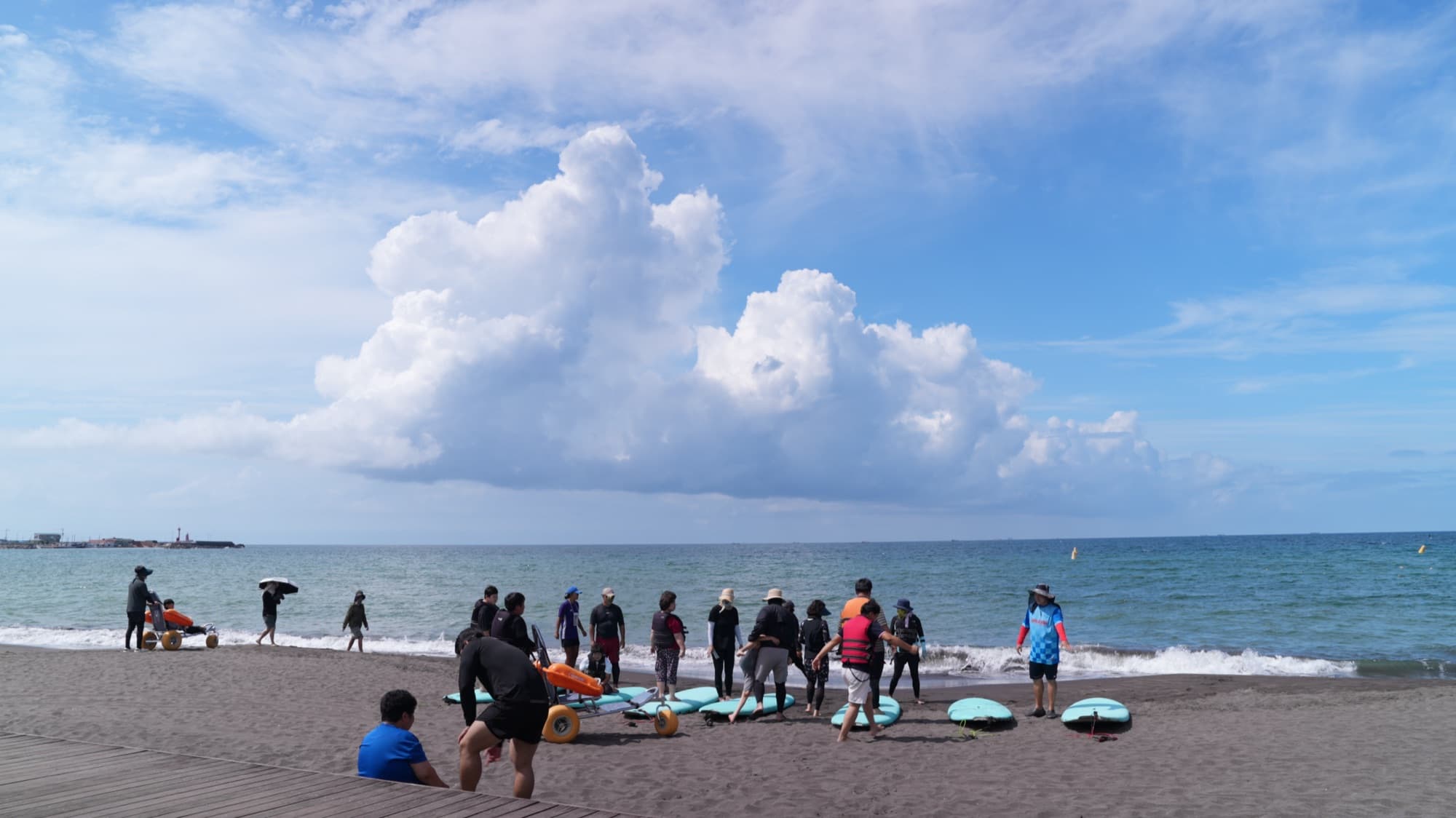 해수욕장 모래사장에 민트색 해변 서프보드가 줄지어 놓여 있고, 구명조끼를 입은 참가자와 자원봉사자들이 보드 곁에 모여 바다를 바라보는 모습. 왼쪽에 노란 벌룬 타이어가 달린 해변용 휠체어가 있고 하늘에 큰 뭉게구름이 떠 있다.