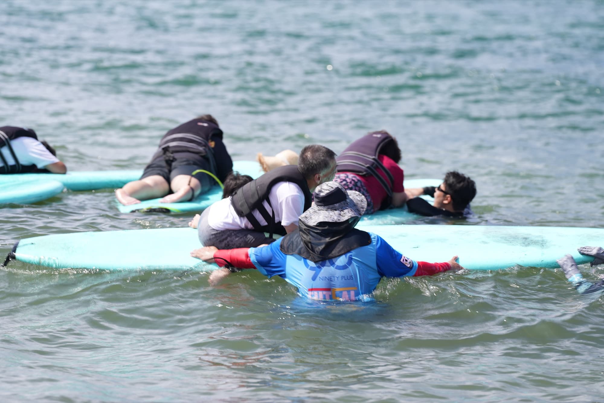 얕은 바다에서 여러 참가자가 민트색 서프보드 위에 엎드려 있고, 파란 래시가드를 입은 강사들이 물속에서 보드를 잡아 안정시키며 파도를 기다리는 모습.